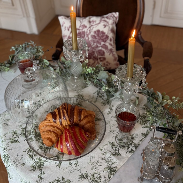 Plato de cristal con croissant y pastel sobre mesa con mantel blanco, dos velas doradas en candelabros de cristal