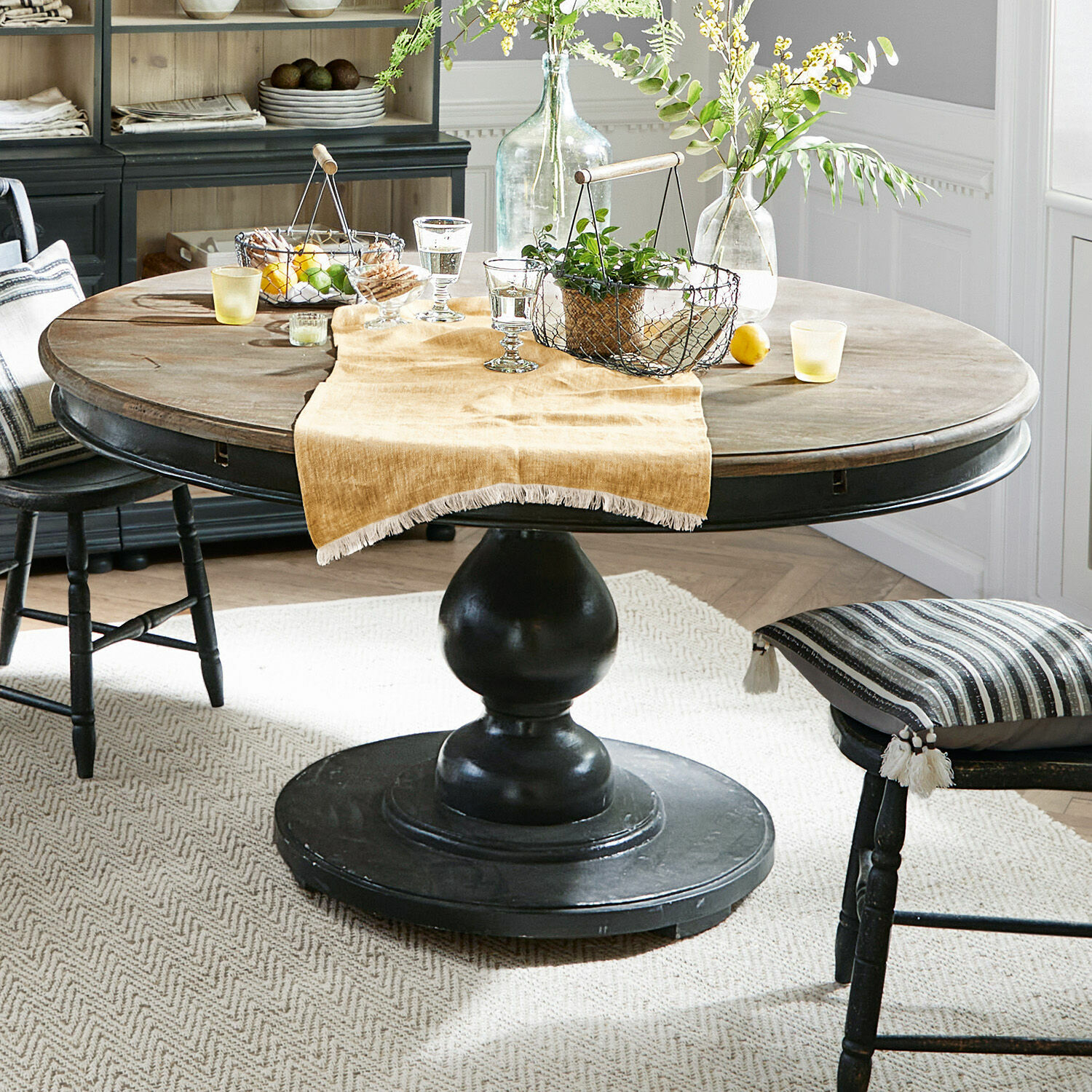 Mesa Shrewsbury Mesa redonda de madera con base negra y decoración sobre mantel beige en comedor