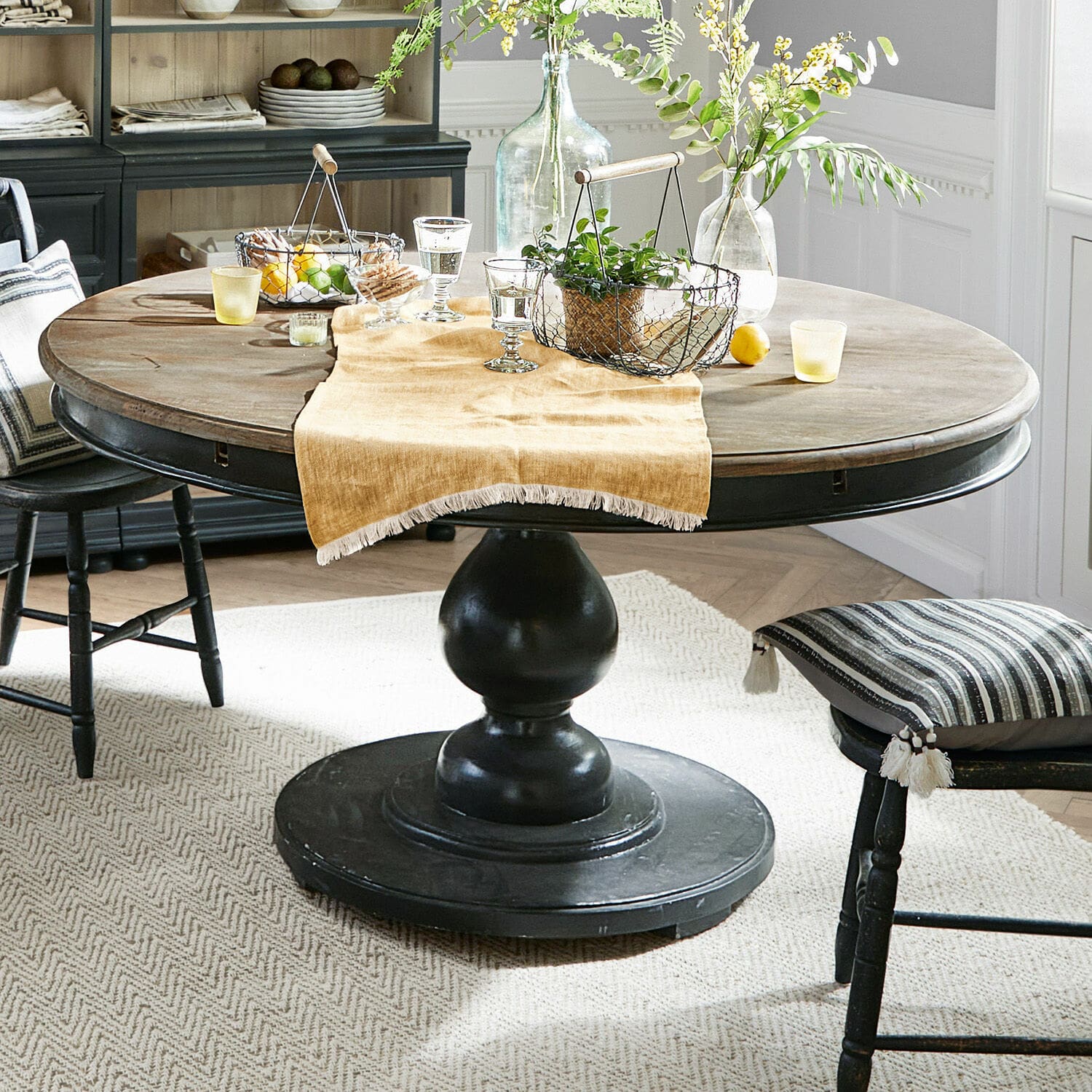 Mesa Shrewsbury Mesa redonda de madera con base negra y decoración sobre mantel beige en comedor