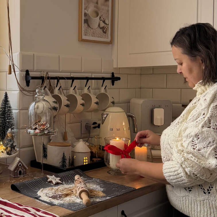 Mujer con suéter blanco decorando vela con lazo rojo en cocina con encimera de madera y decoración navideña