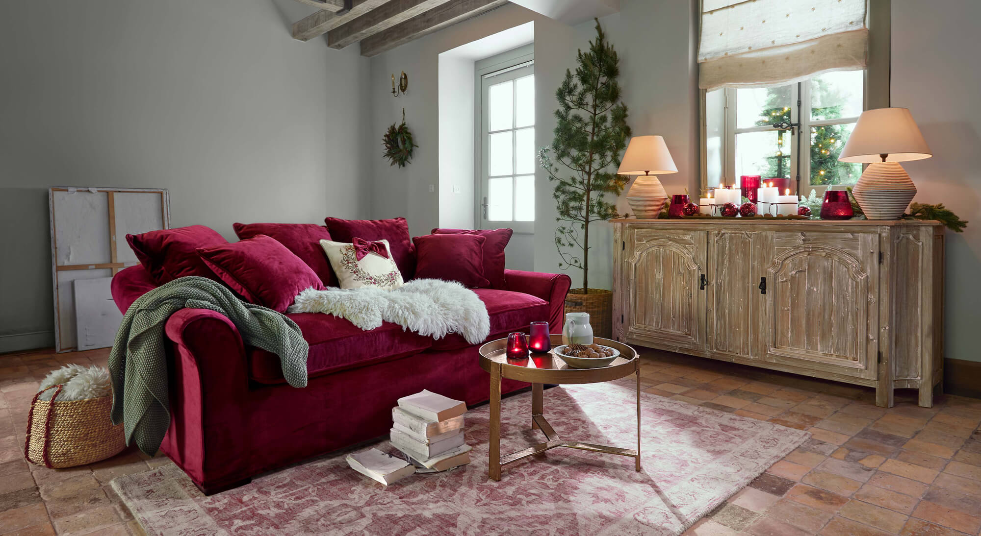 Sofá de terciopelo rojo con cojines y manta verde en salón acogedor con aparador de madera y decoración navideña
