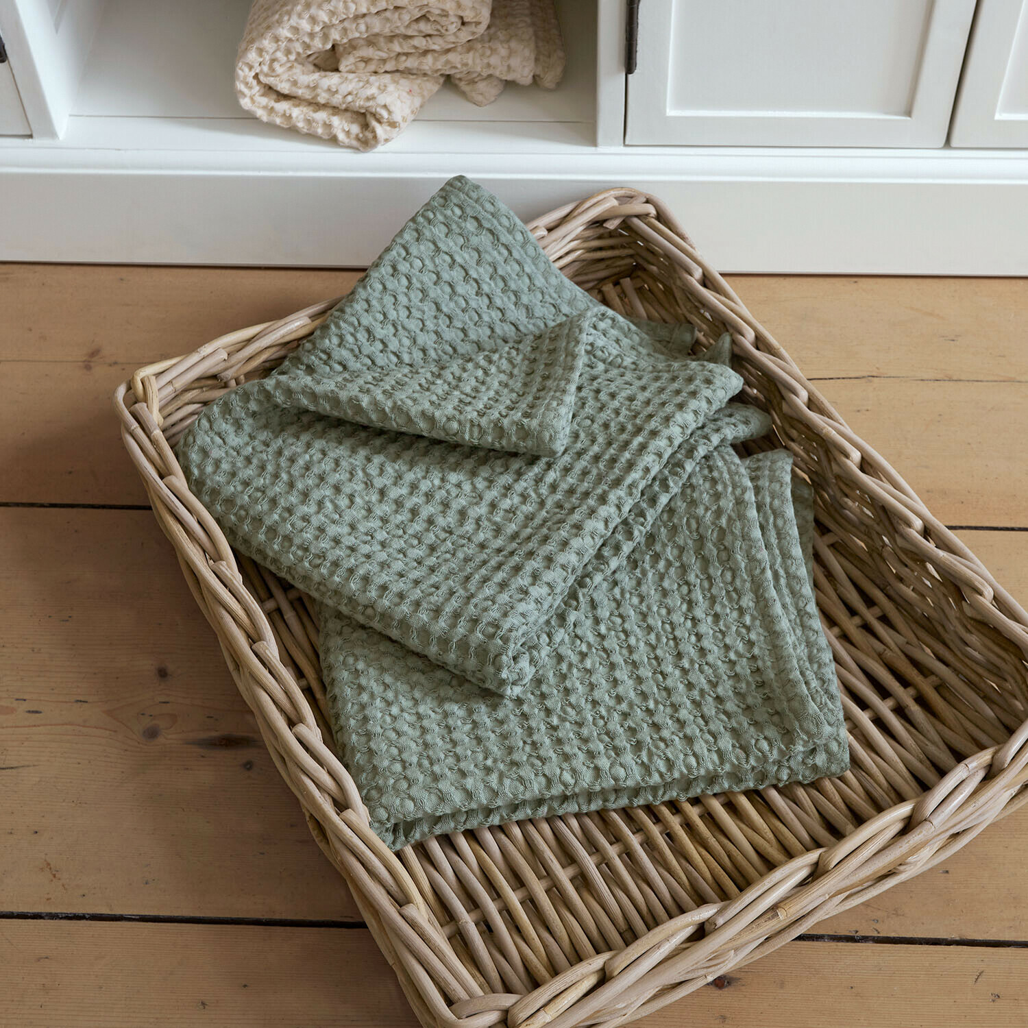 Set de 2 toallas Maralei Manta verde de algodón con textura de gofre en cesta de mimbre sobre suelo de madera