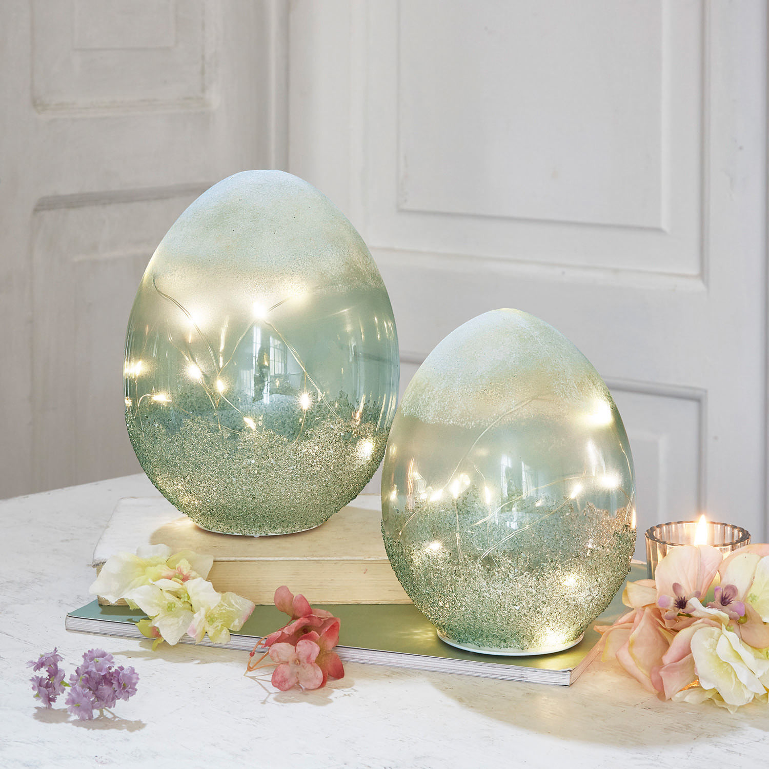 Set de 2 huevos LED Borleos Huevos decorativos de vidrio verde con luces LED integradas sobre mesa con flores y vela