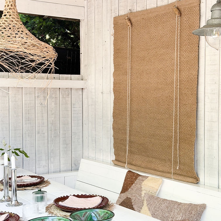 Pared de madera blanca con estor de yute natural y mesa decorada con cojines estilo bohemio