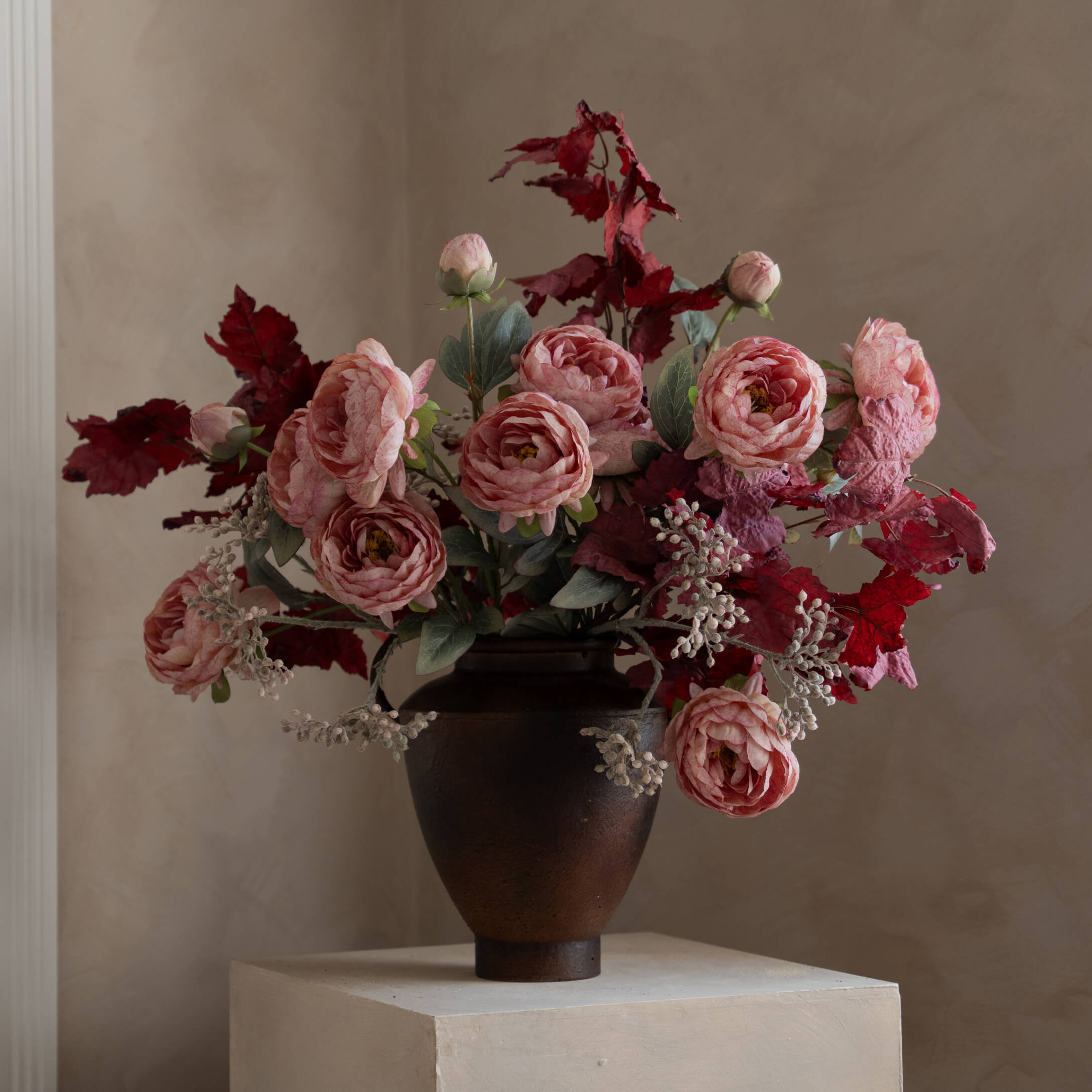 Ramo de rosas rosas y hojas rojas en jarrón cerámico oscuro sobre pedestal claro