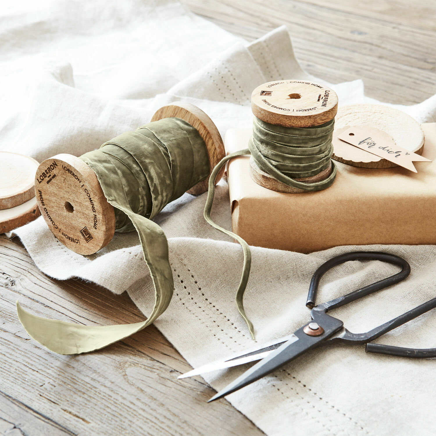 Cinta de terciopelo verde en carretes de madera con tijeras y regalo sobre mantel de lino