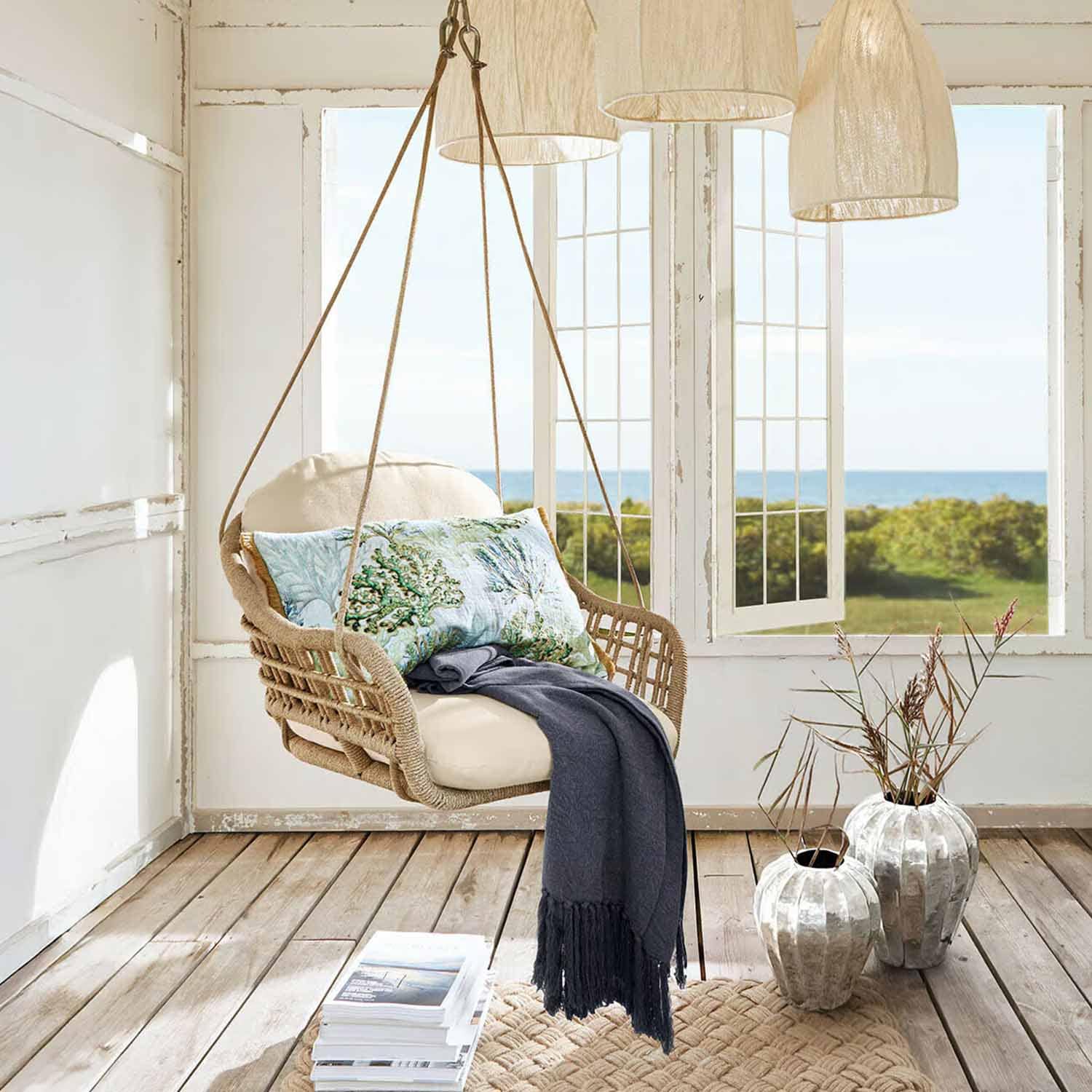 Sillón colgante de ratán con cojín crema y manta azul en habitación luminosa con vistas al mar