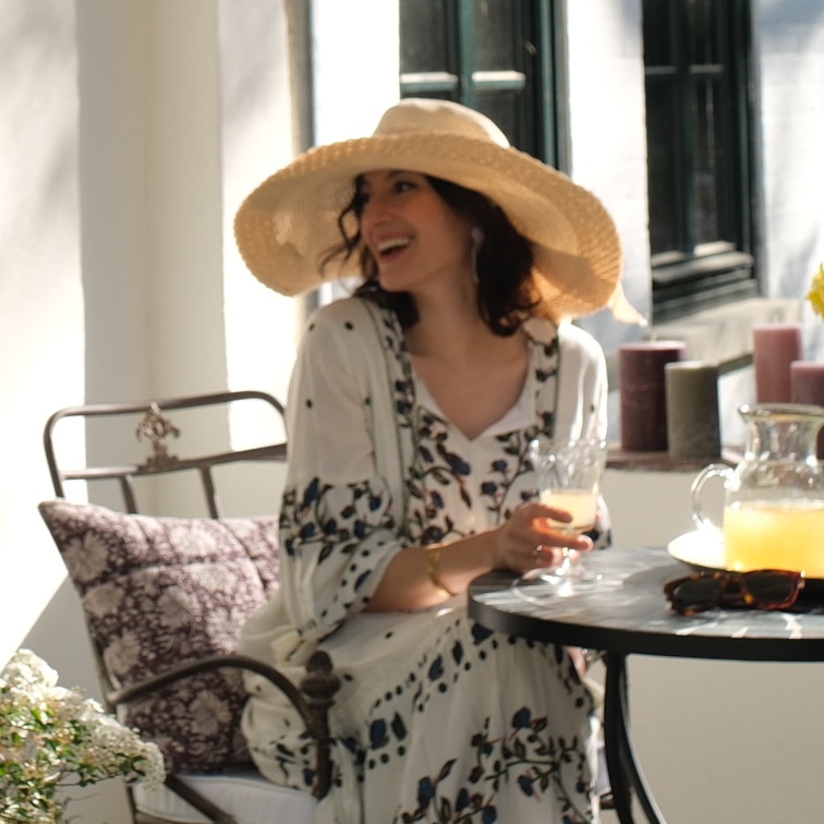 Mujer con vestido floral y sombrero de paja sentada en mesa con bebida en interior luminoso