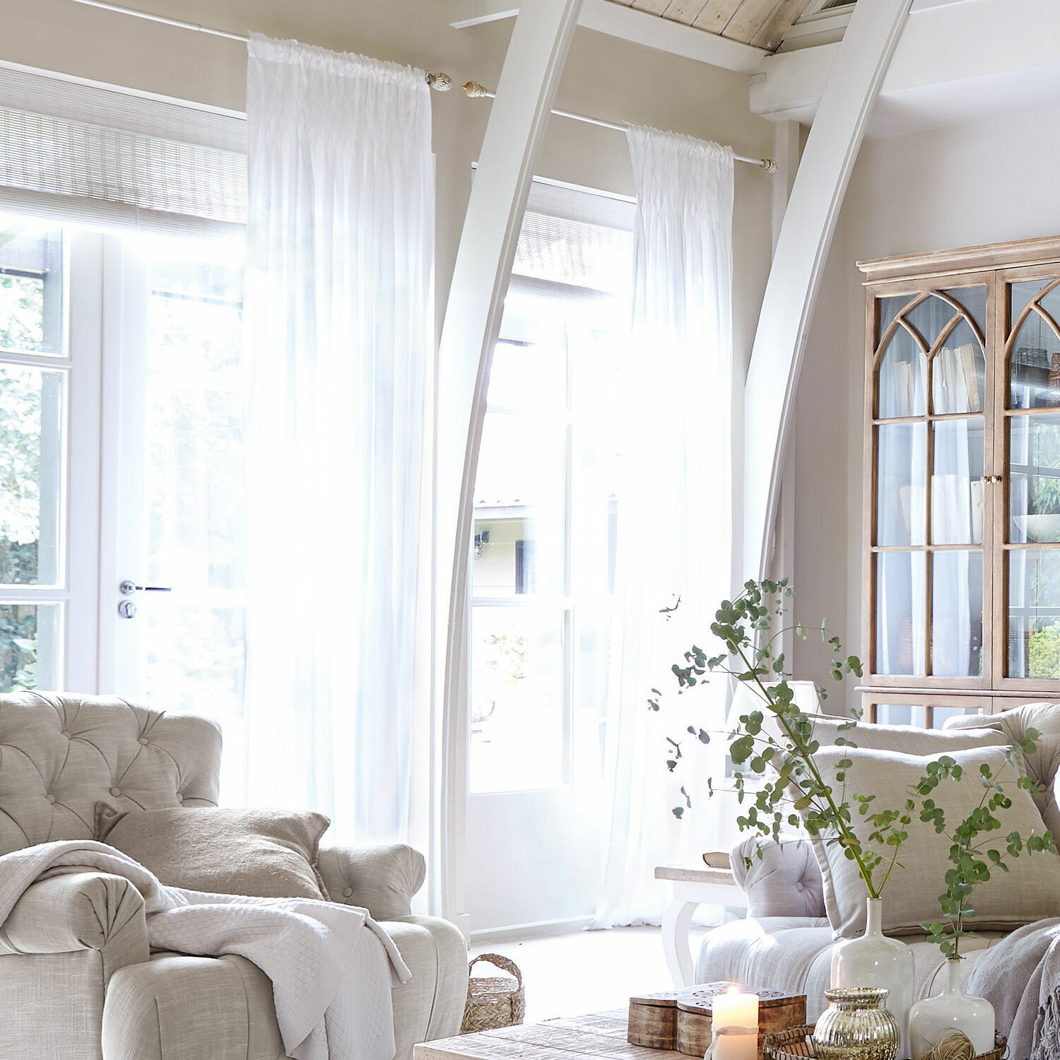 Cortina Otilie Sala luminosa con sillón beige, cortinas blancas y vitrina de madera estilo vintage