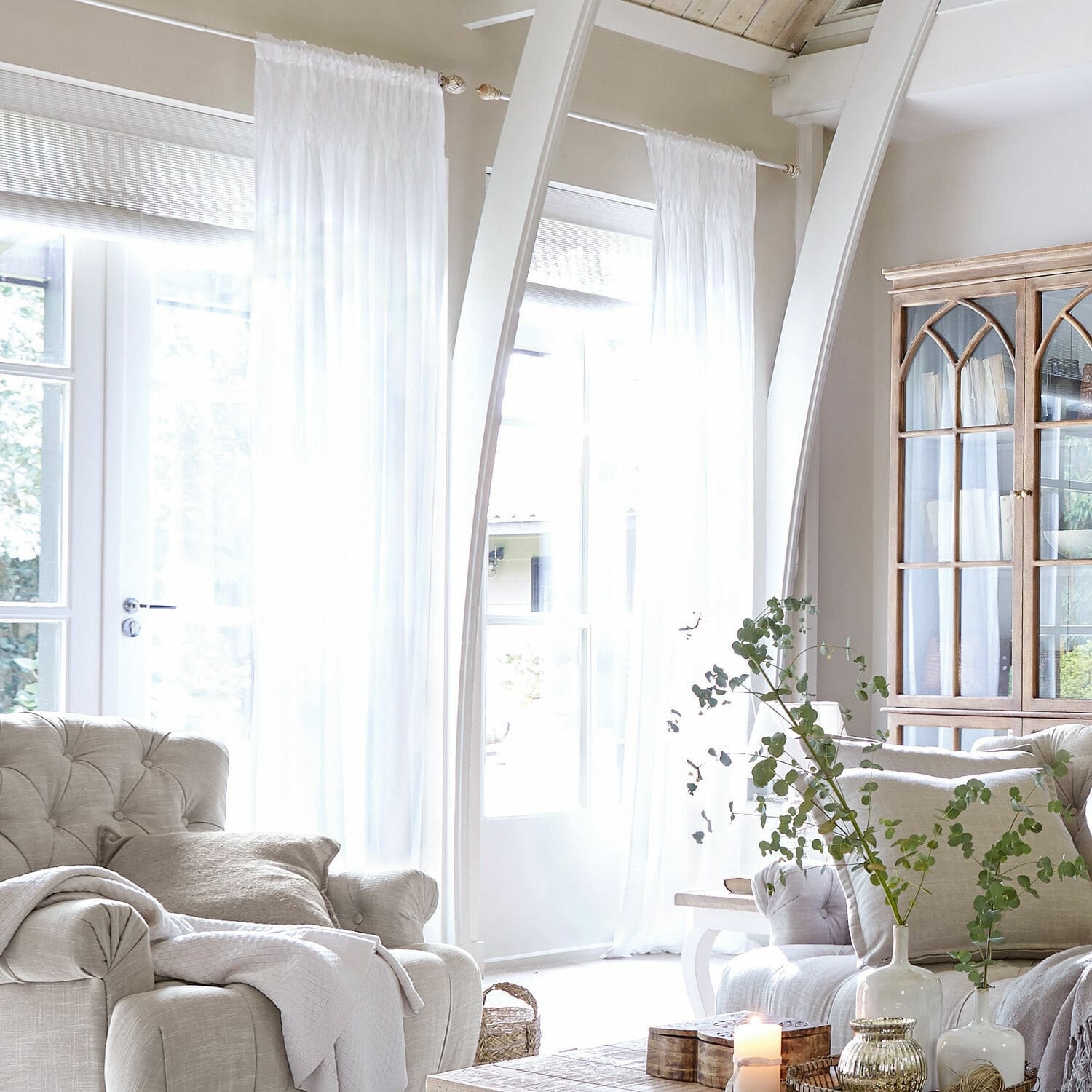 Cortina Otilie Sala luminosa con sillón beige, cortinas blancas y vitrina de madera estilo vintage