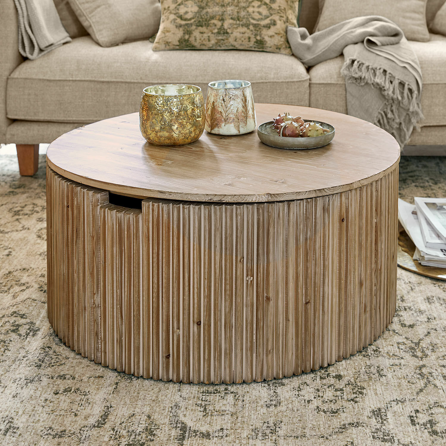 Mesa de centro redonda de madera natural sobre alfombra en sala con sofá beige y mantas