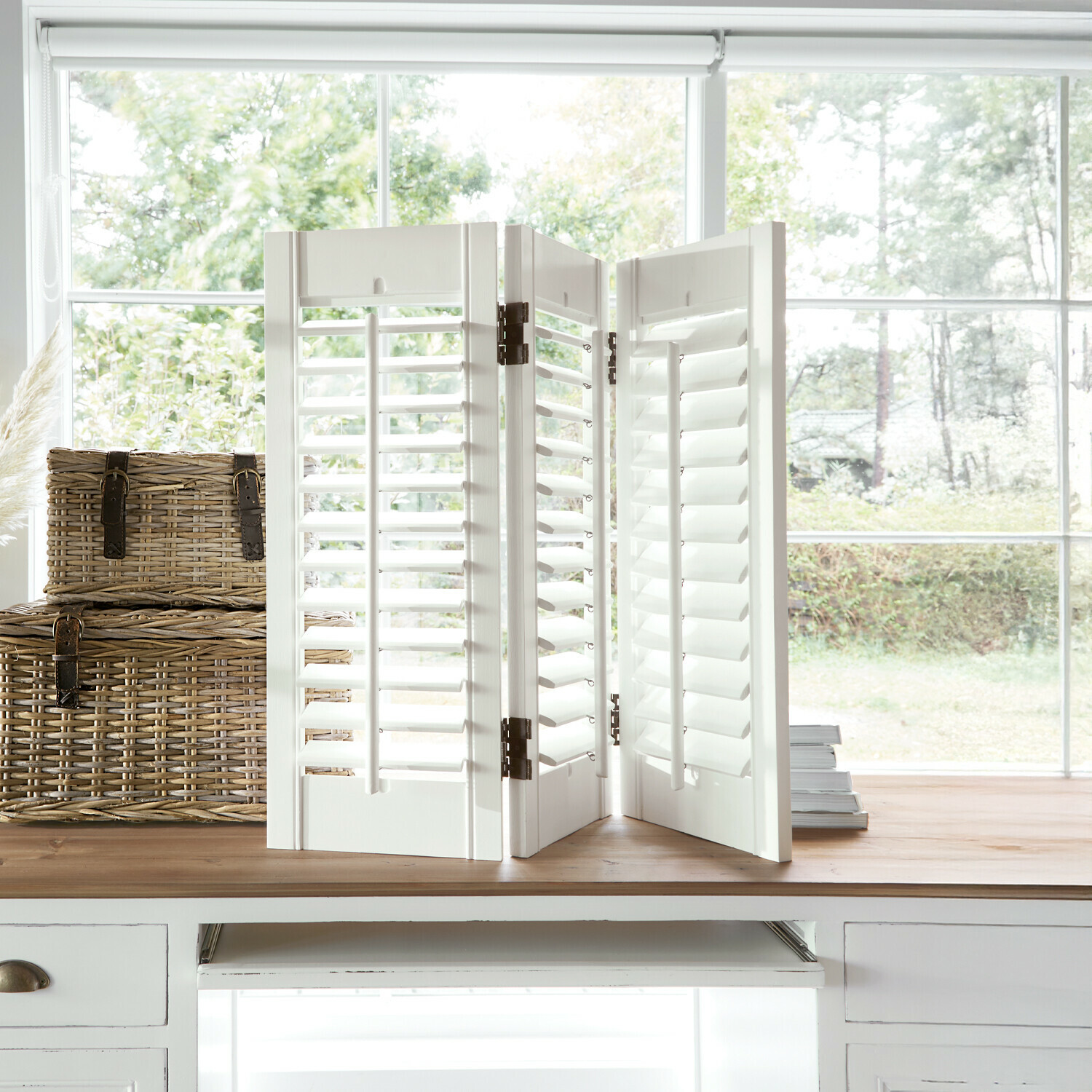 Biombo de madera blanca con lamas móviles sobre mesa de madera junto a ventana, decoración