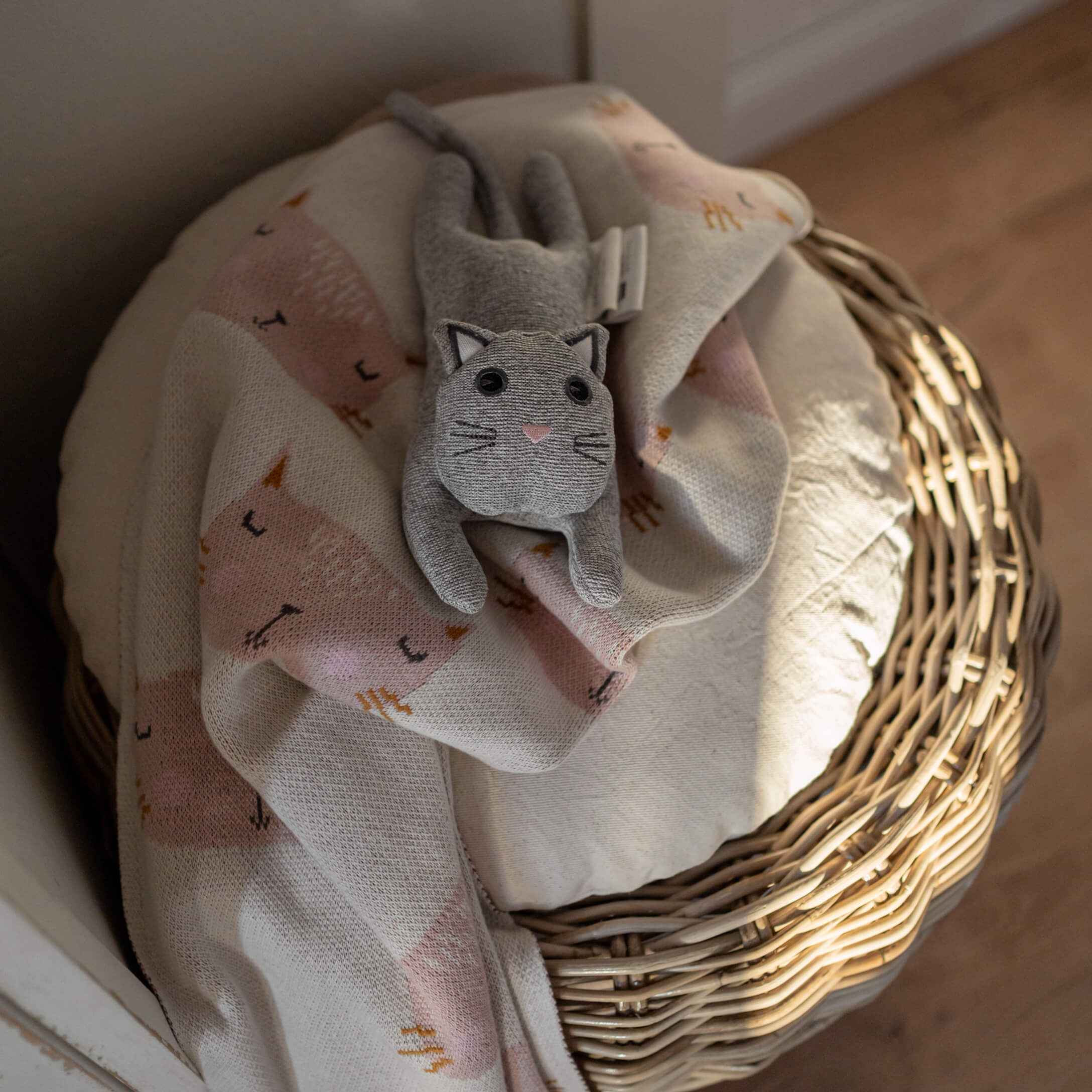 Gato de peluche gris sobre manta rosa claro con diseño de gato en cesta de mimbre