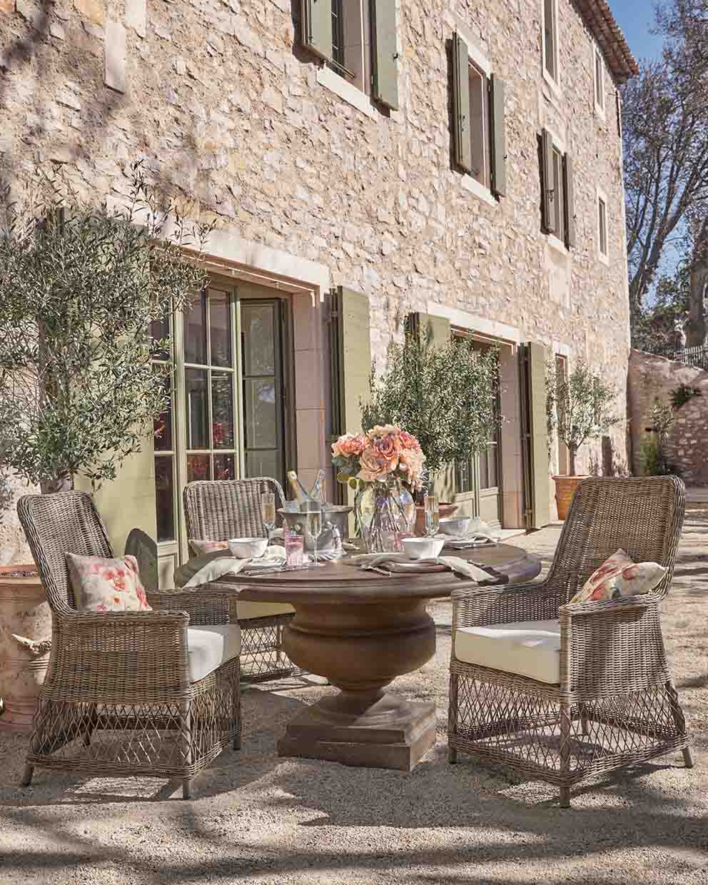 Mesa redonda de piedra con cuatro sillas de mimbre y cojines beige en jardín soleado