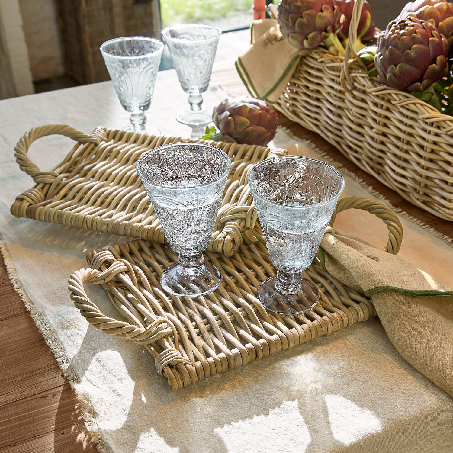 Set de 2 manteles individuales Allegretta Dos vasos decorados sobre bandejas de mimbre en mantel de lino con cesta de alcachofas