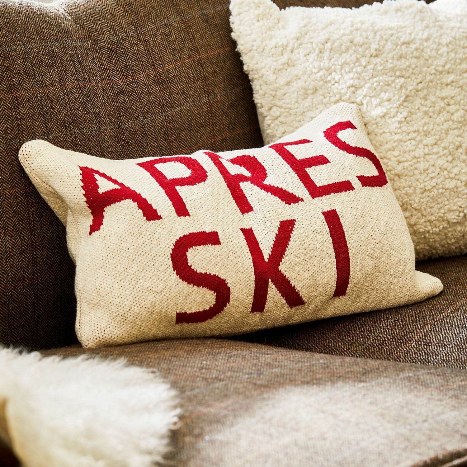 Funda de cojín Apres Ski Cojín beige con texto rojo Apres Ski sobre sofá marrón con manta de piel suave