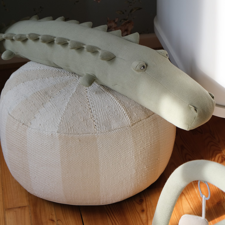 Cocodrilo de peluche verde claro sobre un puf beige tejido en suelo de madera, decoración infantil