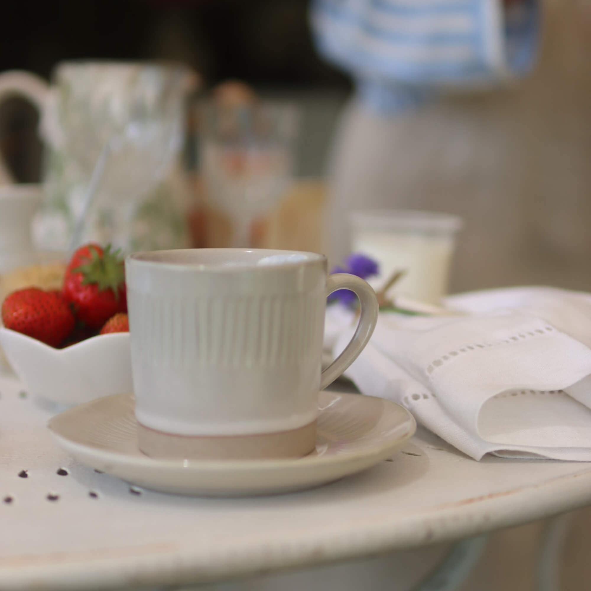 Taza de cerámica beige con plato sobre mesa blanca con fresas y servilleta de lino
