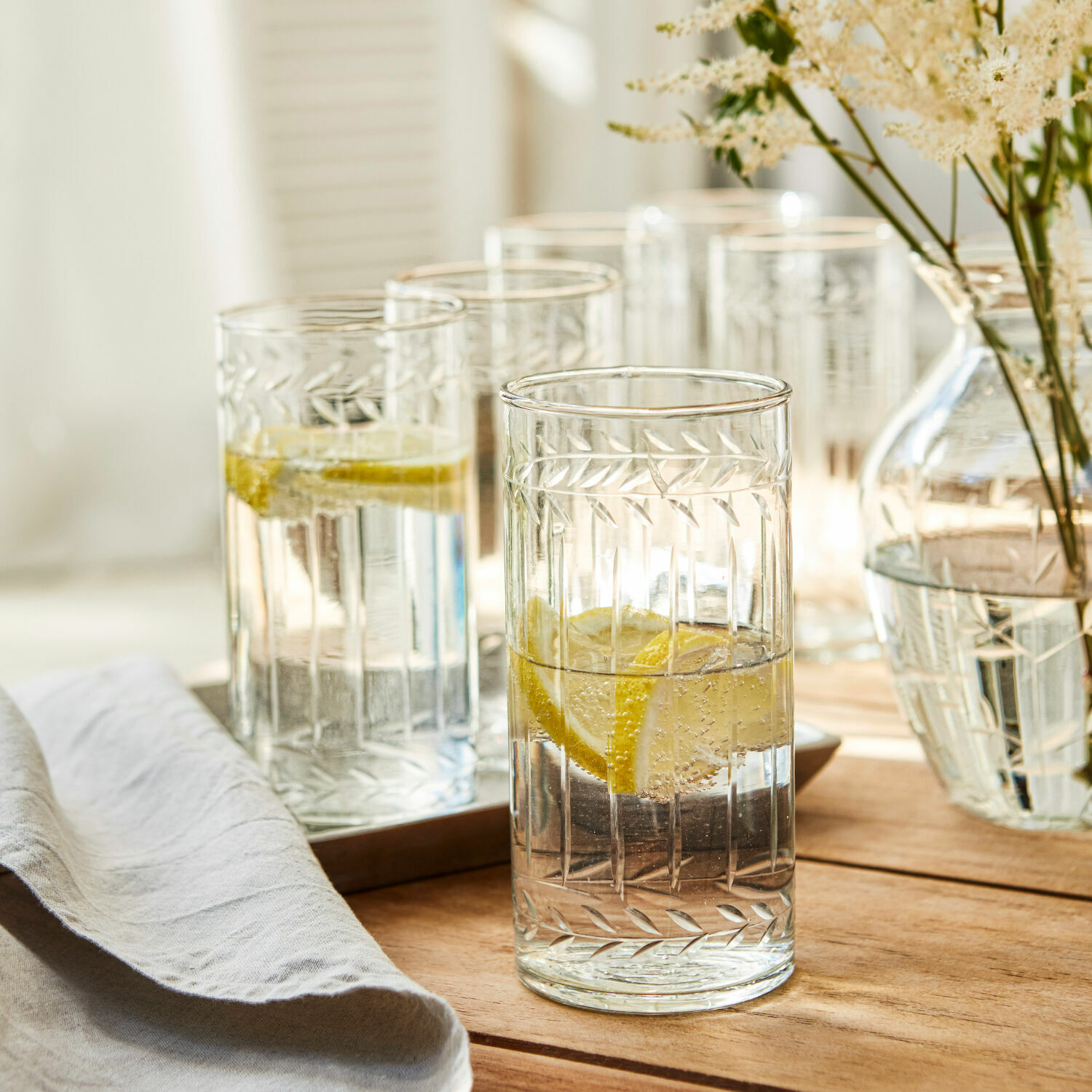 Vasos de vidrio transparente con diseño grabado de hojas, llenos de agua con limón sobre mesa de madera