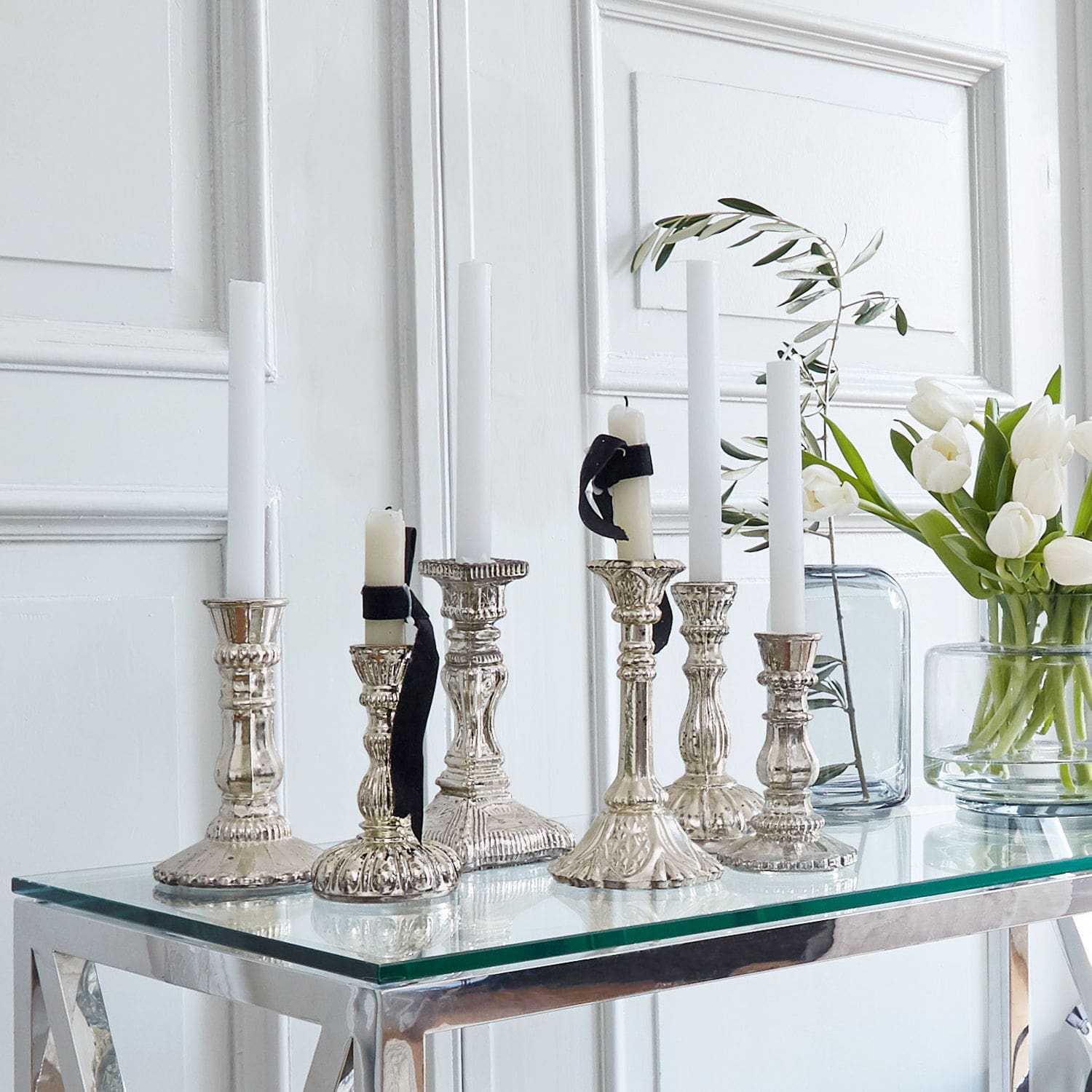 Set de 2 candeleros Adolpha Conjunto de candelabros metálicos plateados con velas blancas sobre mesa de cristal en interior luminoso