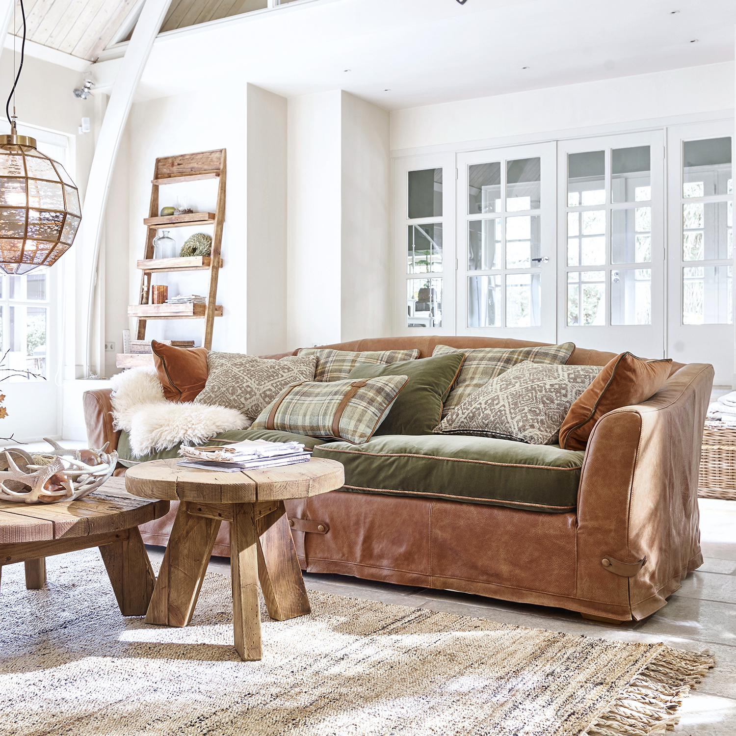 Sofá de cuero marrón con cojines verdes y estampados en salón luminoso con muebles rústicos de madera