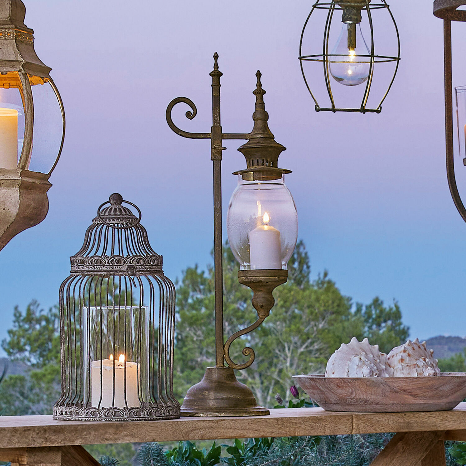 Farol Bowral Linterna metálica con vidrio transparente y vela encendida sobre mesa de madera con decoración de conchas al aire libre