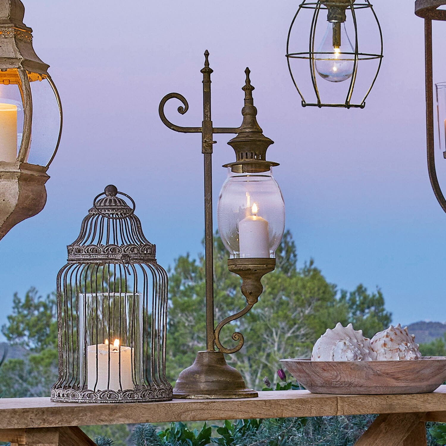 Farol Bowral Linterna metálica con vidrio transparente y vela encendida sobre mesa de madera con decoración de conchas al aire libre