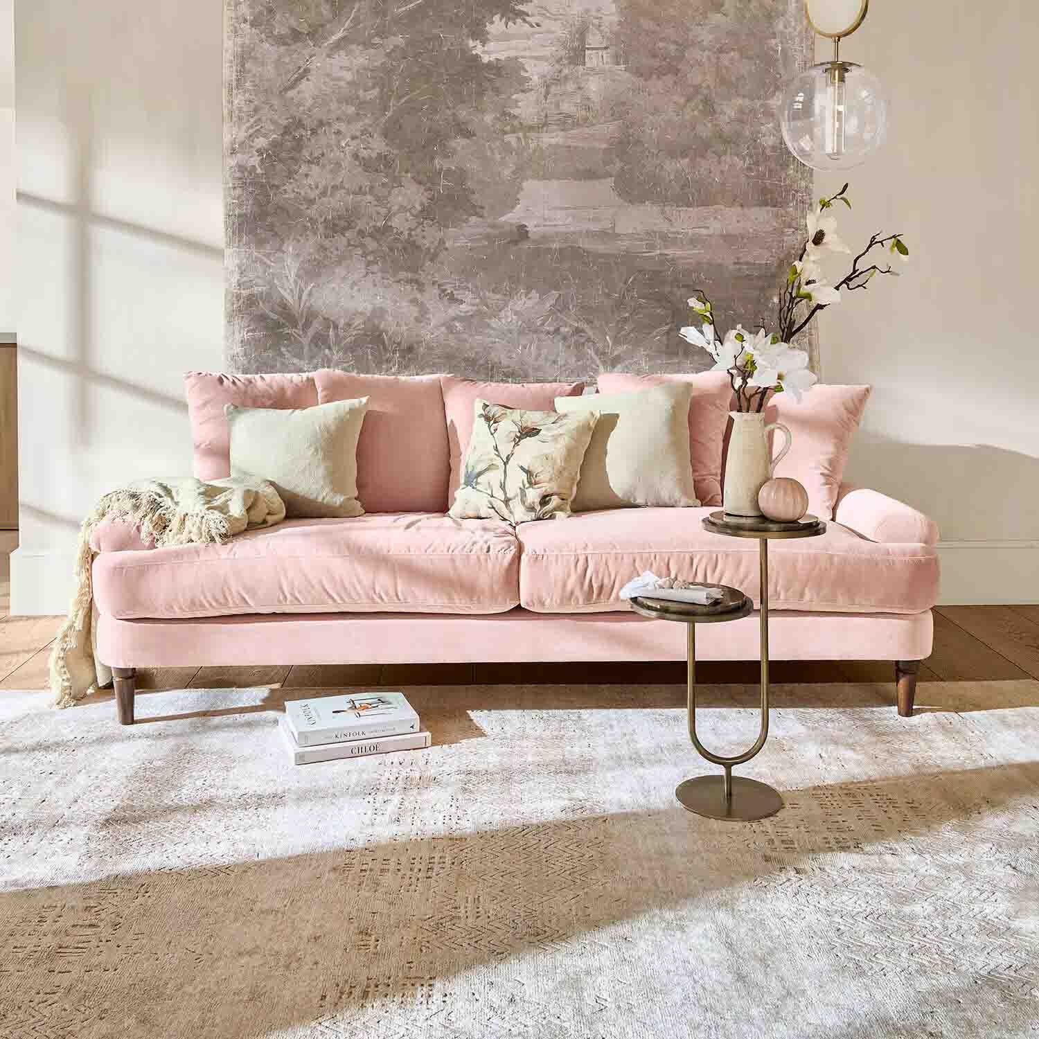 Sofá de terciopelo rosa con cojines y mesa auxiliar en salón luminoso con decoración mural