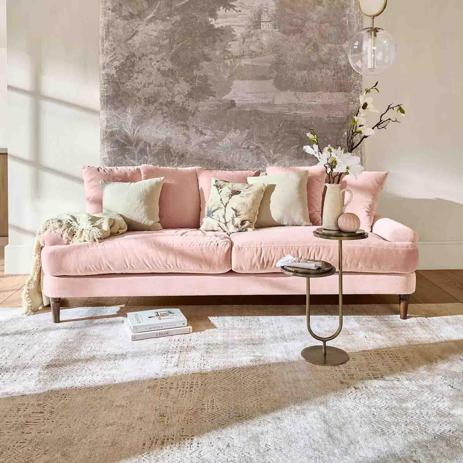Sofá de terciopelo rosa con cojines y mesa auxiliar en salón luminoso con decoración mural