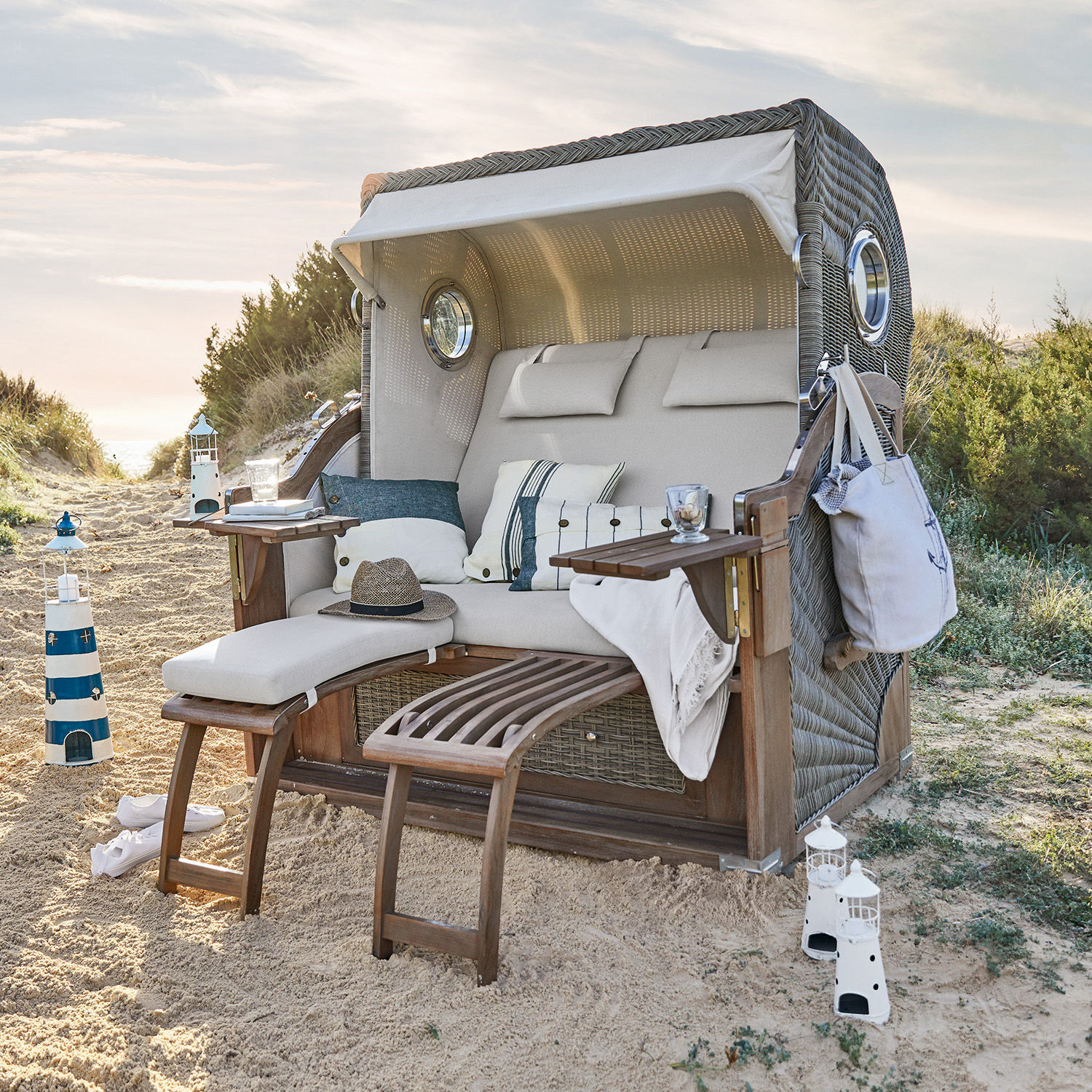 Sillón de playa de mimbre con cojín beige y reposapiés de madera sobre arena, muebles de jardín