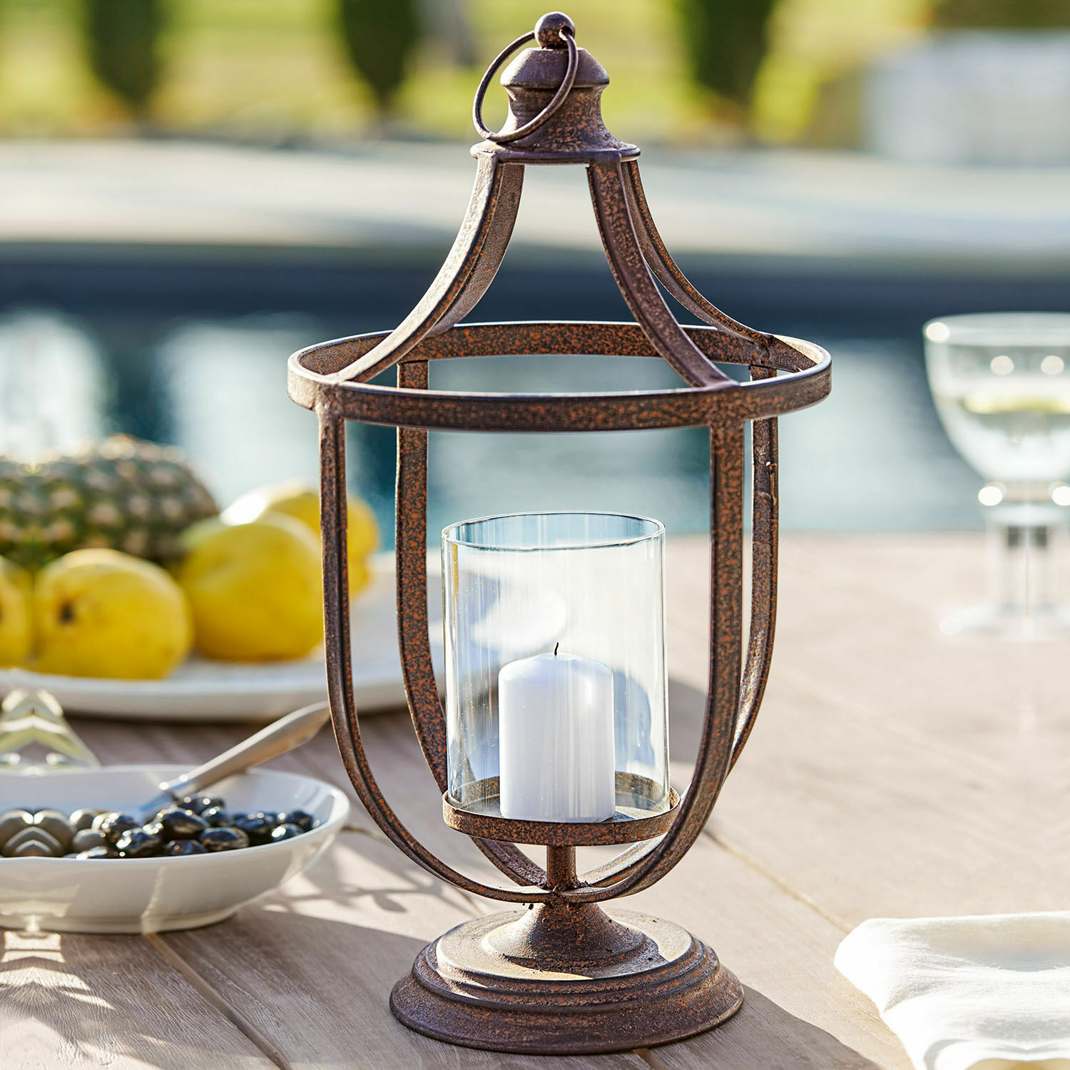 Farol Saint-Denis Linterna decorativa de metal color óxido con vela blanca y vidrio protector sobre mesa de jardín