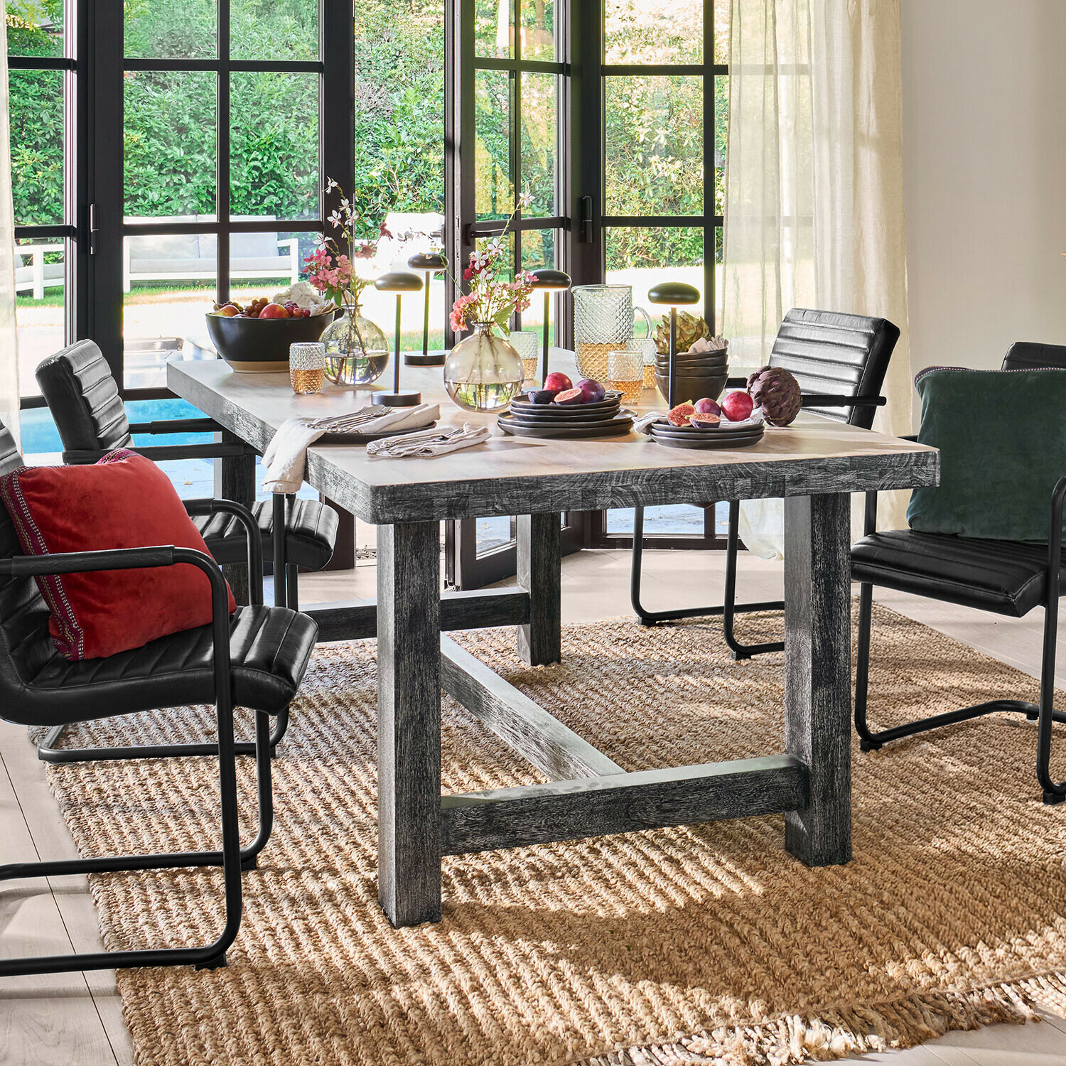 Mesa Oaklynne Mesa de comedor de madera gris con sillas negras y decoración frente a grandes ventanas