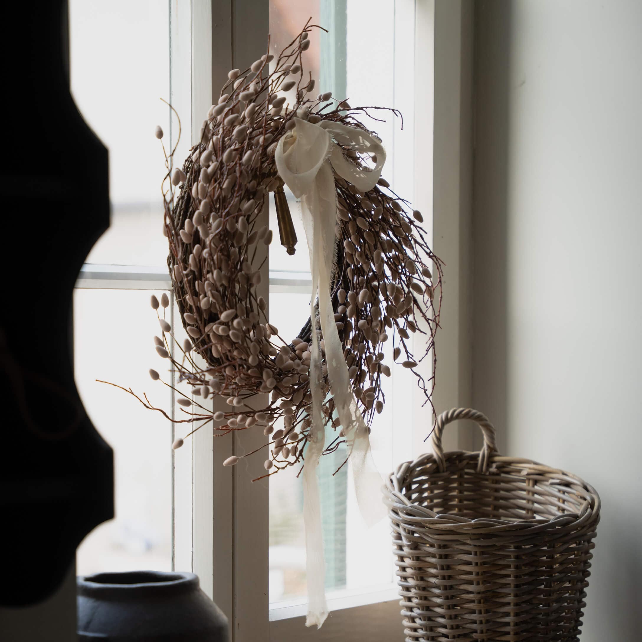 Corona decorativa beige de ramas y capullos con lazo blanco colgada en la ventana, cesta de mimbre al lado