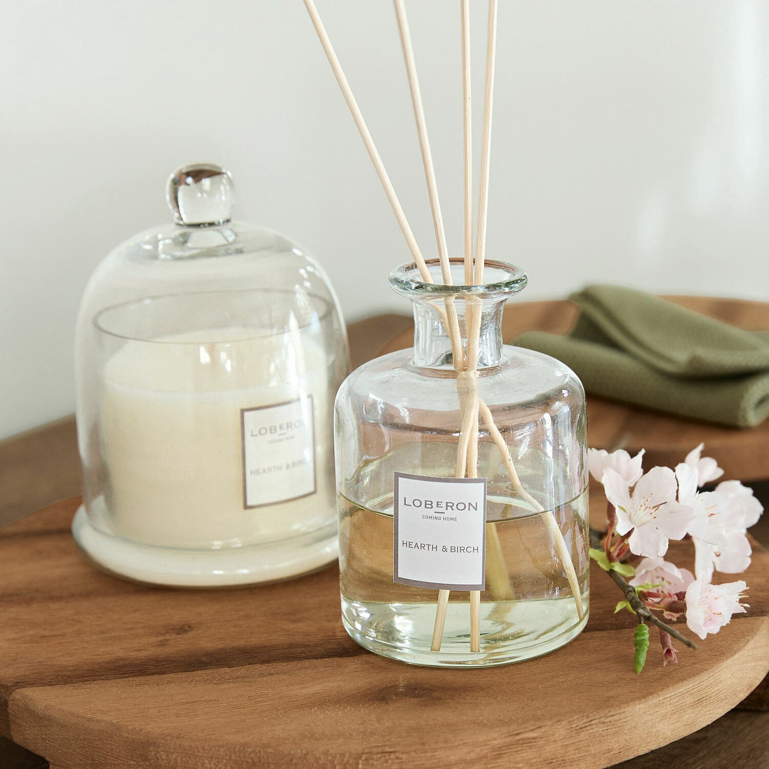Set de fragancias Vanis Difusor de aroma de vidrio Hearth & Birch con varillas sobre mesa de madera y vela blanca bajo campana