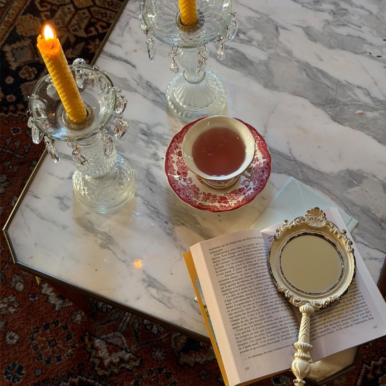 Candelabros de cristal con velas amarillas y taza con diseño rojo sobre mesa de mármol con libro y espejo de mano