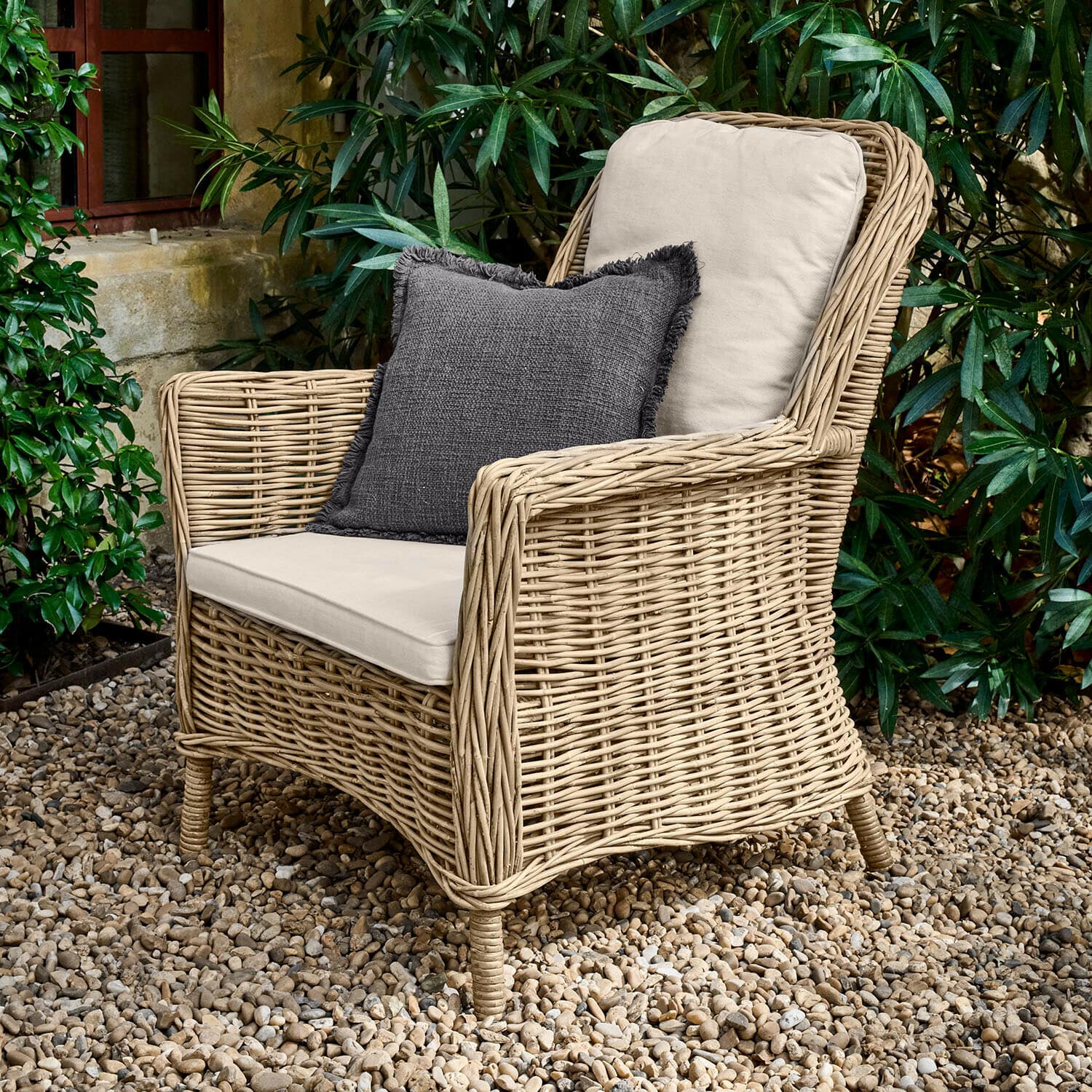 Sillón Travenne Sillón de ratán con cojines crema y cojín decorativo gris oscuro en patio de grava