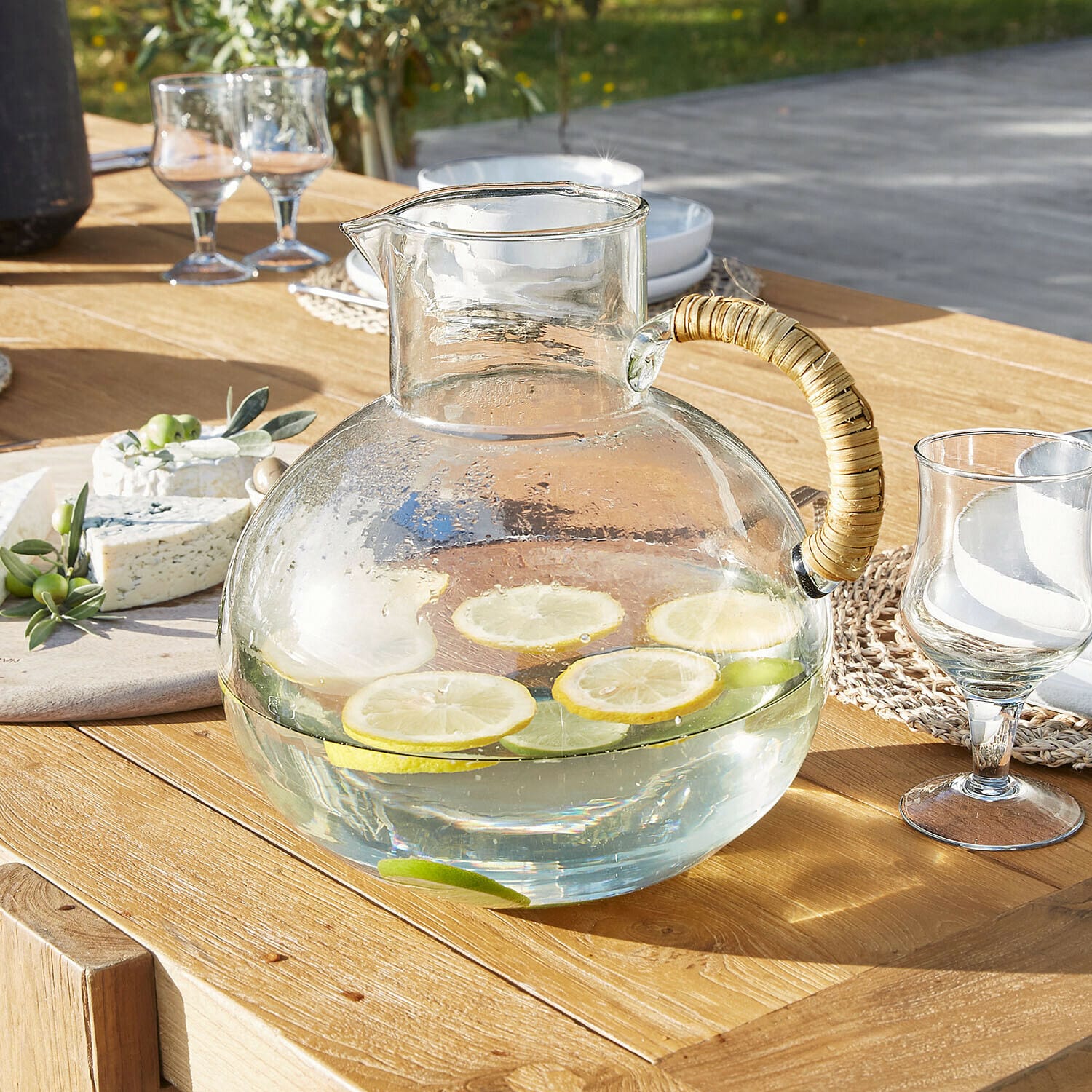 Jarra Stormea Jarra de vidrio con asa de ratán sobre mesa de madera, agua con rodajas de limón y lima