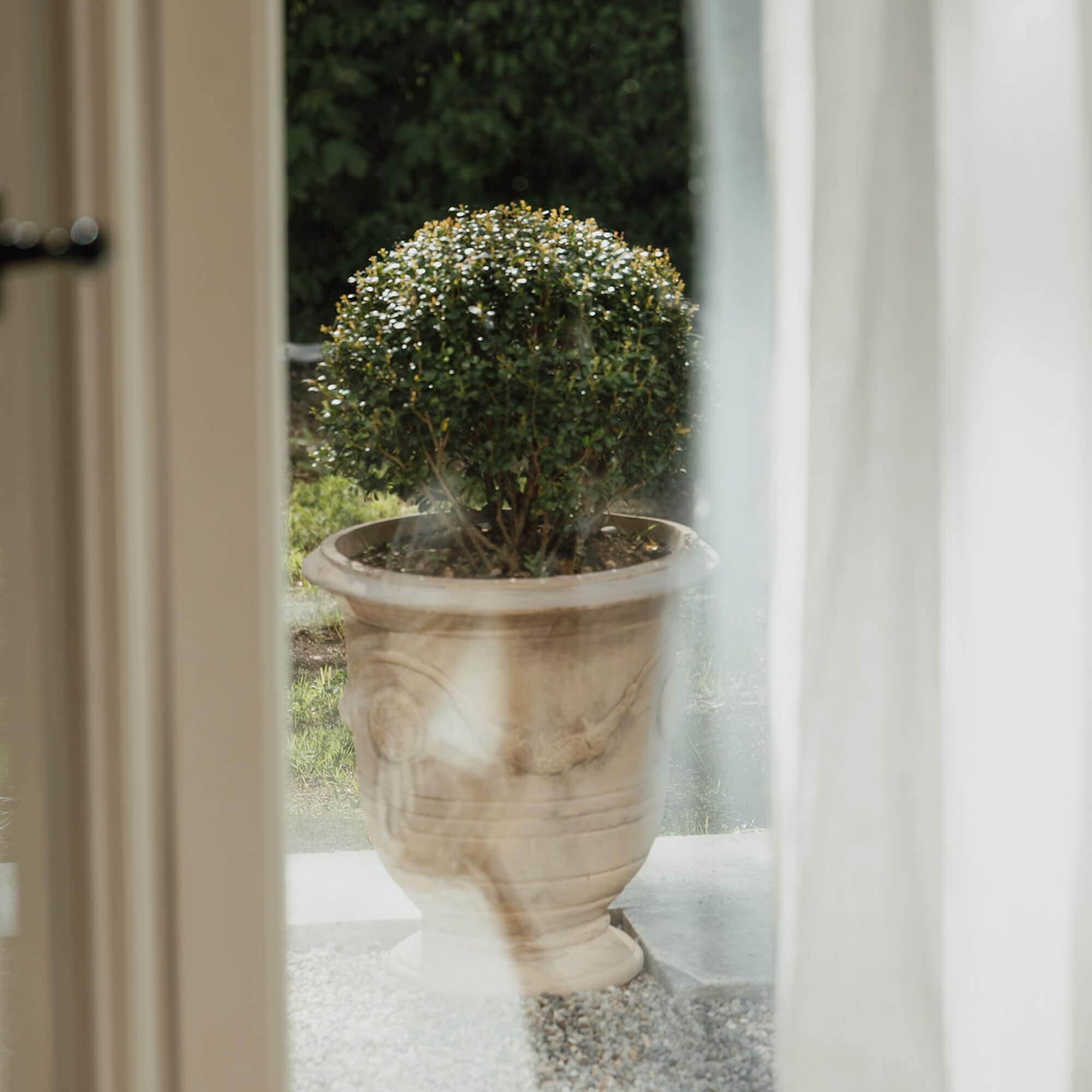 Maceta decorativa de piedra clara con arbusto verde frente a una casa