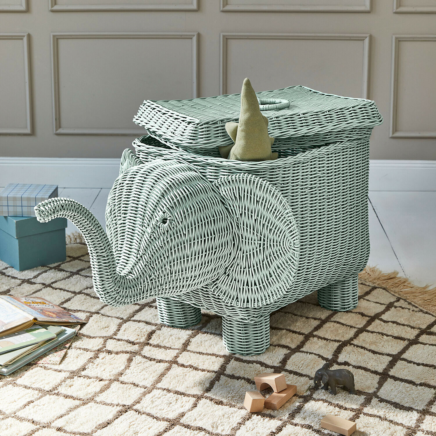 Cesta de ratán verde menta con forma de elefante, tapa abierta y juguetes sobre alfombra