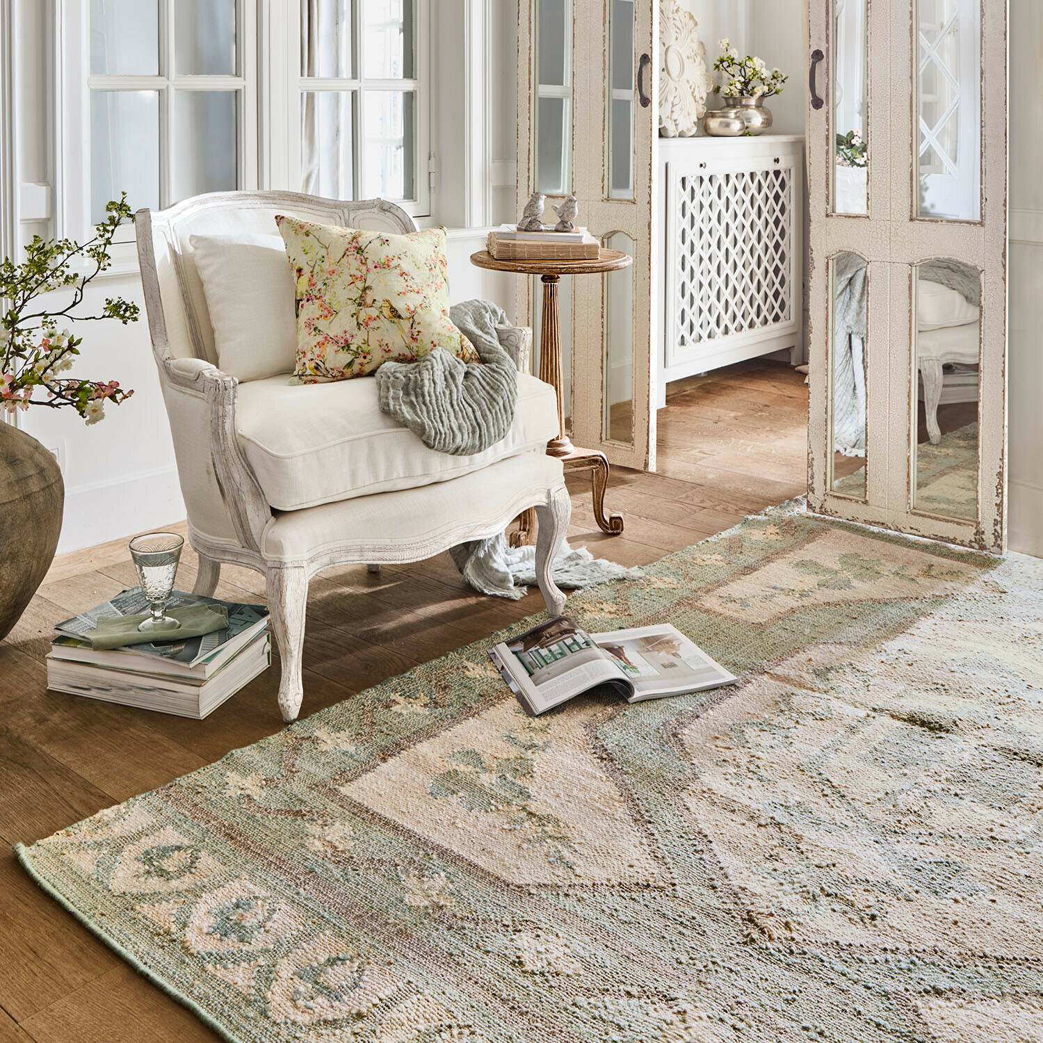 Alfombra Raneema Sillón blanco de madera con cojín floral en sala luminosa con alfombra vintage en tonos suaves