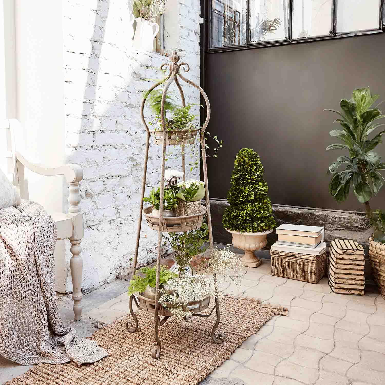 Macetero de metal de pie con tres niveles y plantas verdes en interior luminoso con pared de ladrillo blanco