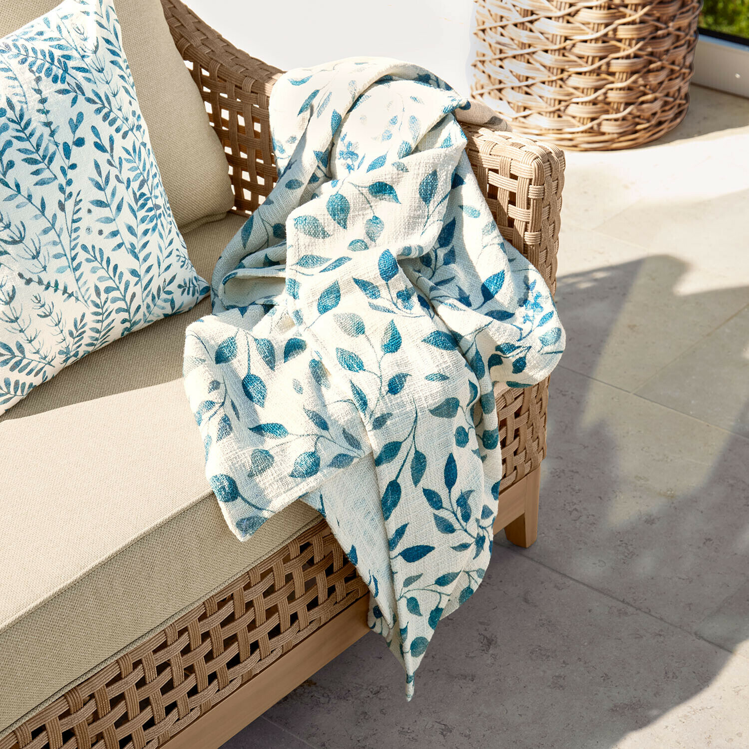 Sofá beige con respaldo trenzado y manta crema con estampado de hojas azules en terraza