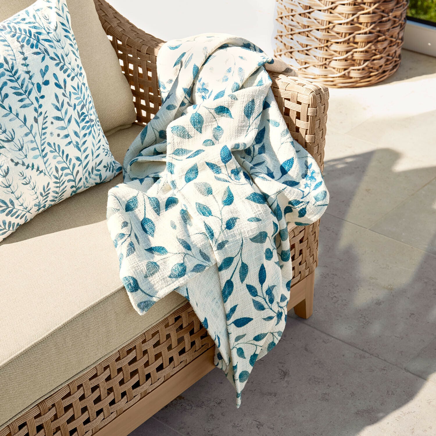Plaid Ocevara Sofá beige con respaldo trenzado y manta crema con estampado de hojas azules en terraza