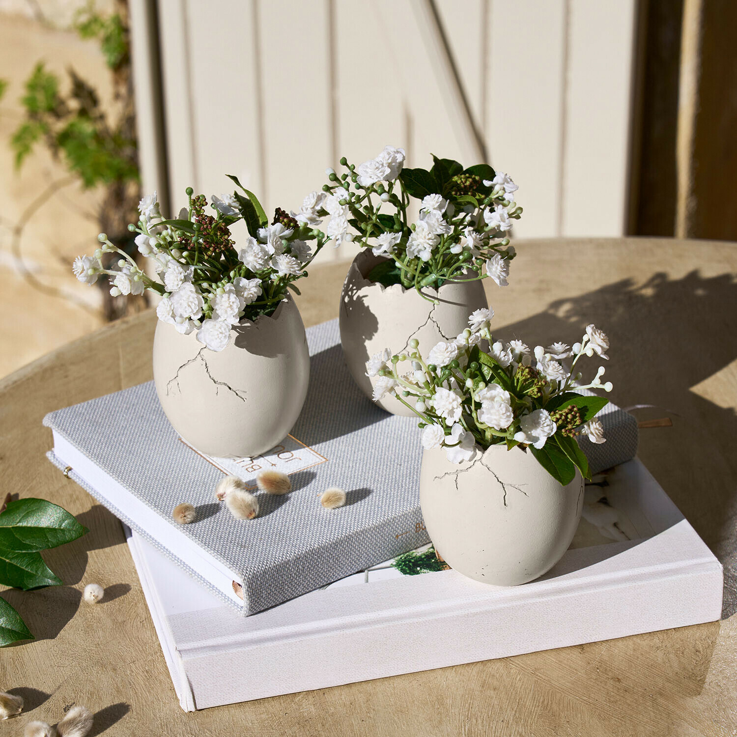 Set de 3 huevos decorativos Waverlye Tres jarrones de cerámica blanca en forma de huevo con flores blancas sobre mesa de madera con libros