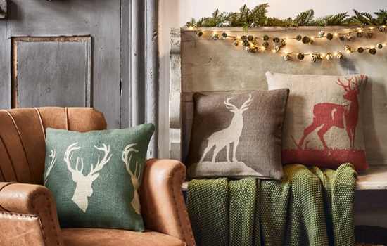 Cojines decorativos con motivo de ciervo en colores naturales sobre sillón de cuero y banco con manta verde