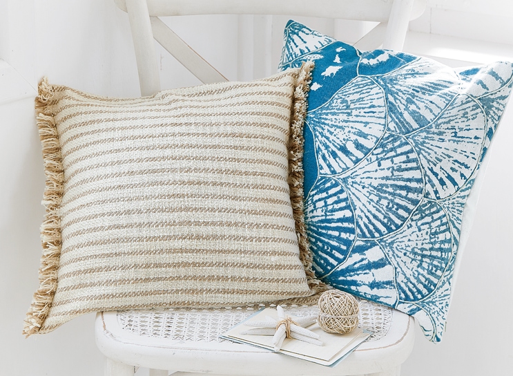 Dos cojines decorativos en silla blanca, uno beige con rayas y flecos, otro azul con diseño de conchas