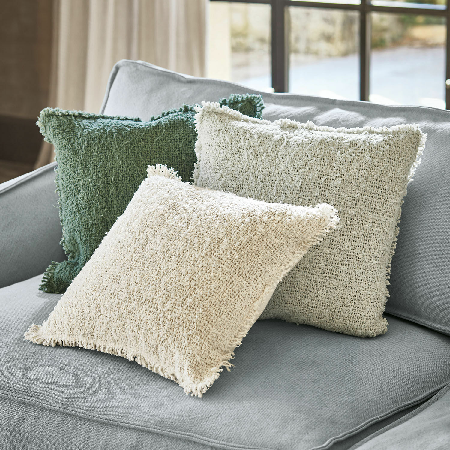 Tres cojines decorativos de tela en tonos crema y verde sobre un sofá gris