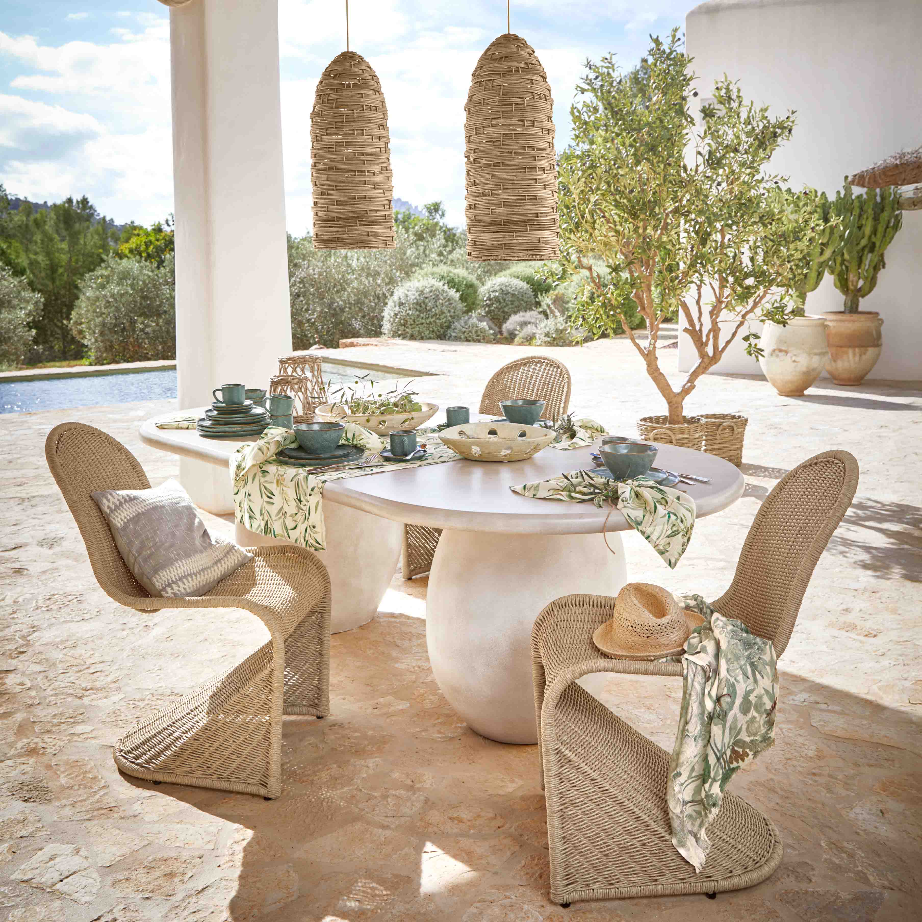 Mesa ovalada de piedra blanca con sillas de mimbre y vajilla verde en exterior junto a la piscina