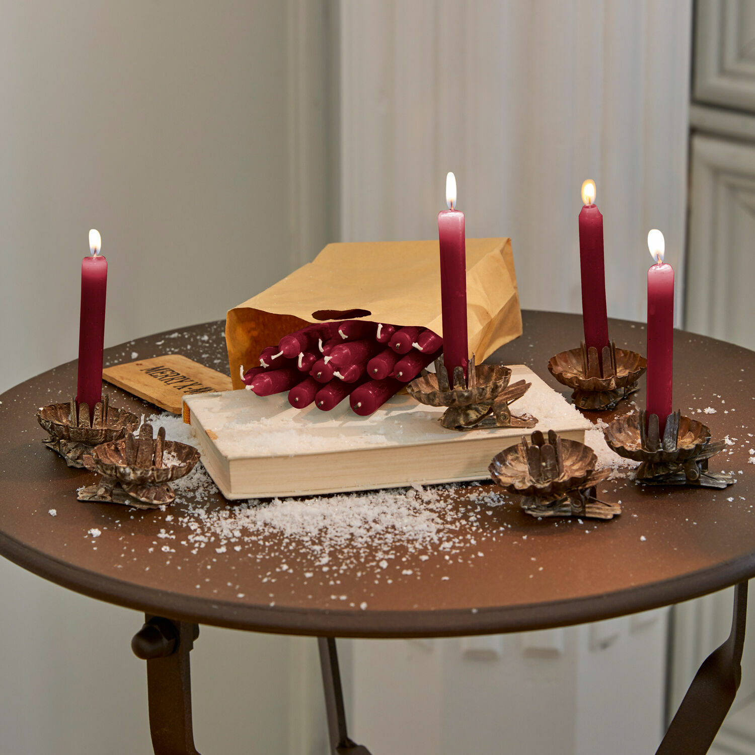 Set de 20 velas Noufa Velas burdeos con pinzas sobre mesa metálica, bolsa de papel y nieve artificial para decoración navideña