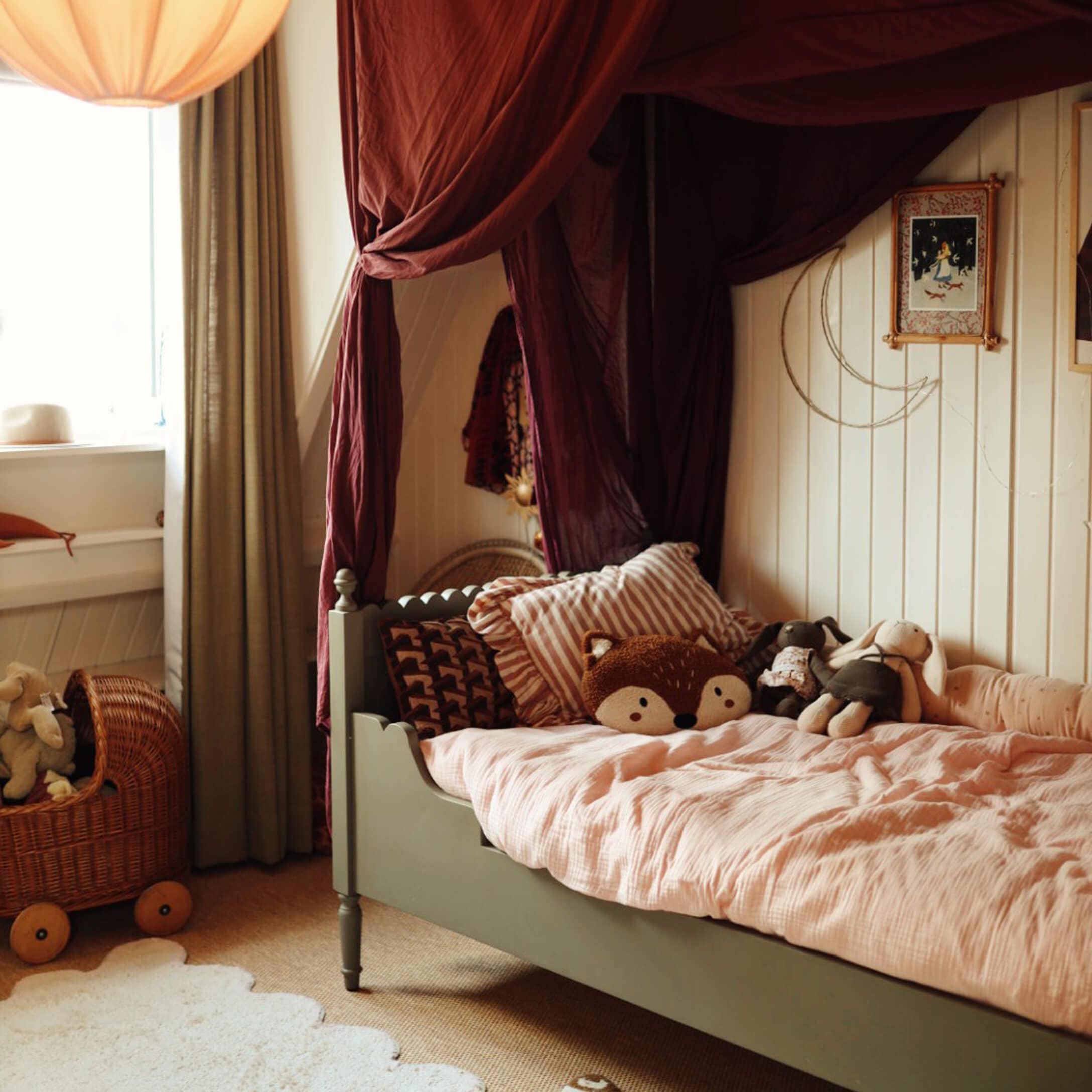 Cama infantil verde con ropa de cama rosa y peluches en habitación cálida con cortinas burdeos